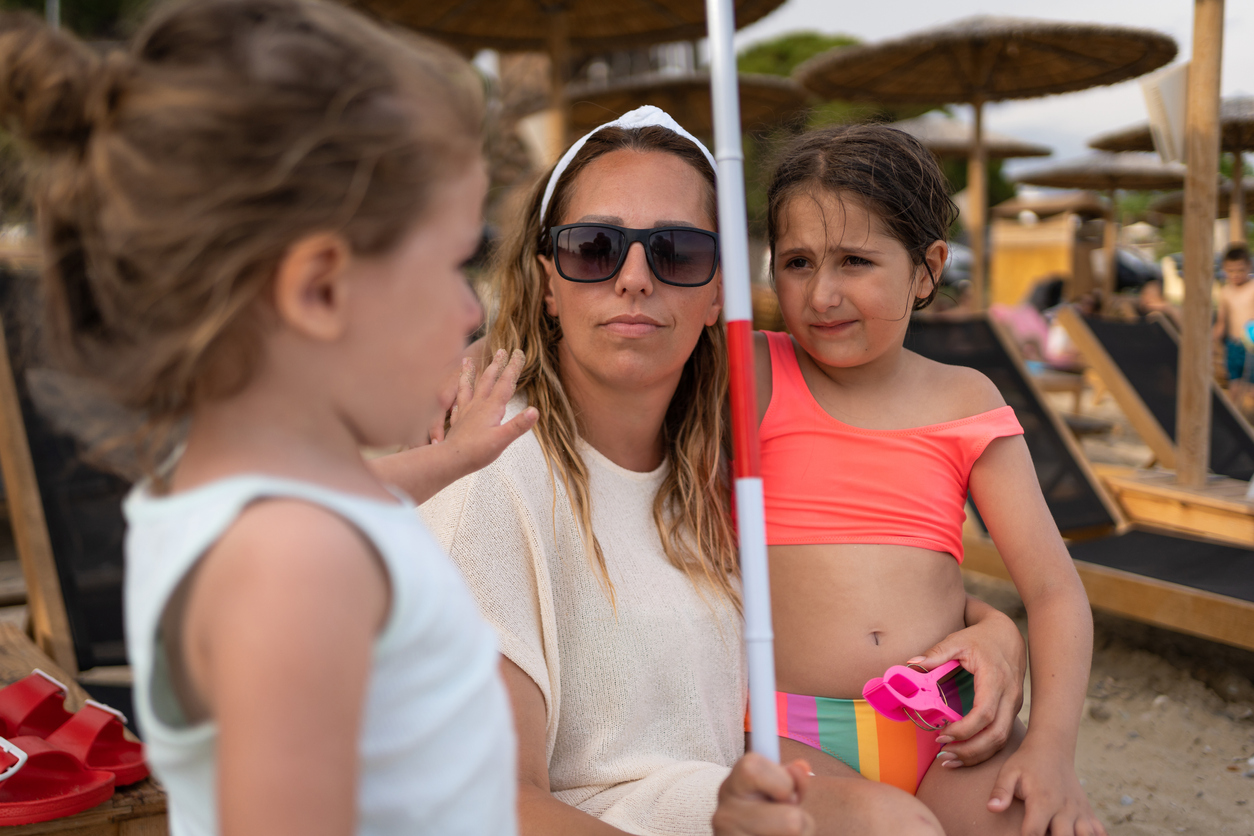 Portrait of Caucasian blind mother, sitting on the beach with walking cone, while consoling her sad daughter