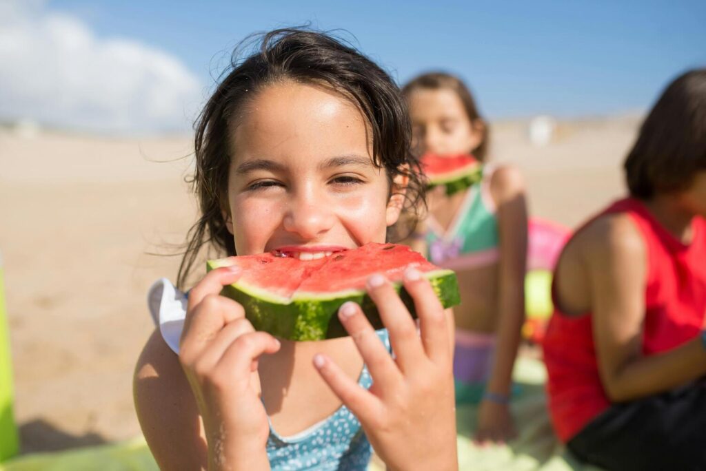 A girl holding and eating a slice of watermelon in a beach sitting on the sand.