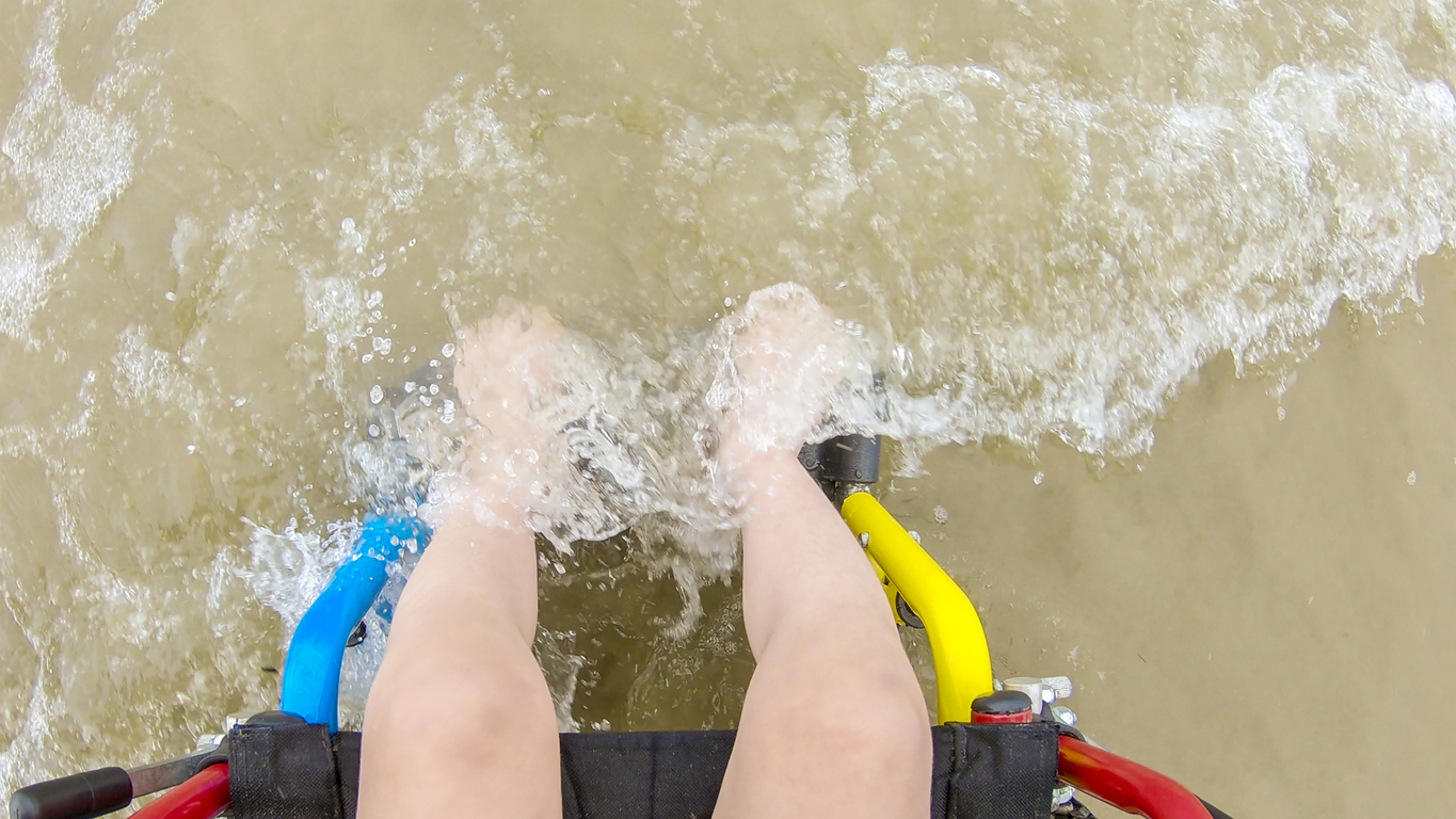 Picture taken from above of a children's legs feeling the water of a wave in the beach. The child is sitting on a wheelchair.