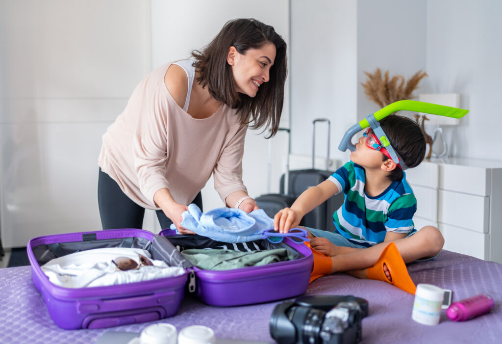 Mother and son packing for a vacation putting clothes away in a purple suitcase laying on a bed.